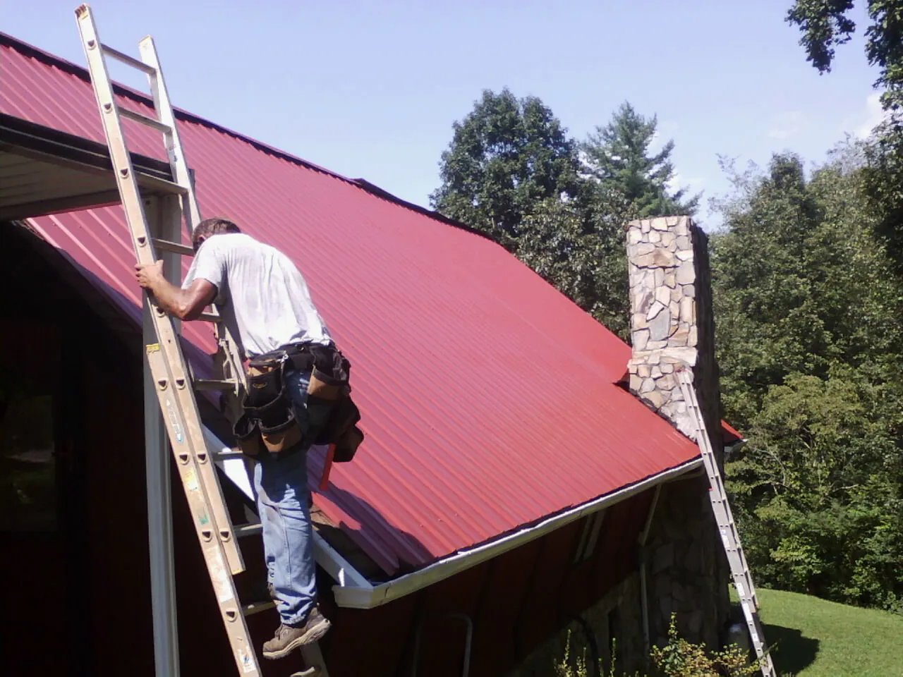 Roofer on a ladder beside a red metal roof and stone chimney — Soffit & Fascia Repair by Roofing Brothers in Durand